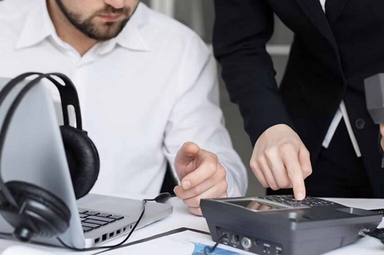 Office workers comparing tasks on a desk phone and laptop, illustrating the difference between Virtual Receptionists vs Automated Phone Service